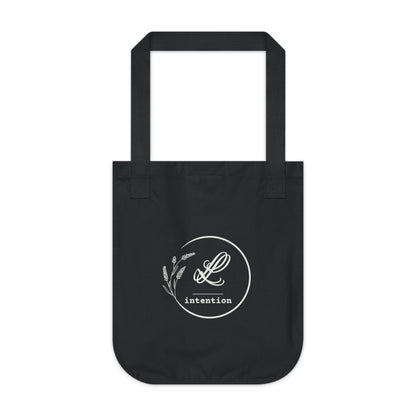Back view of a black canvas tote bag featuring the LL Intention logo: “LL” in cursive above a horizontal line, with the word “intention” in typewriter-style font below, centered within a circular design accented by lavender flowers.