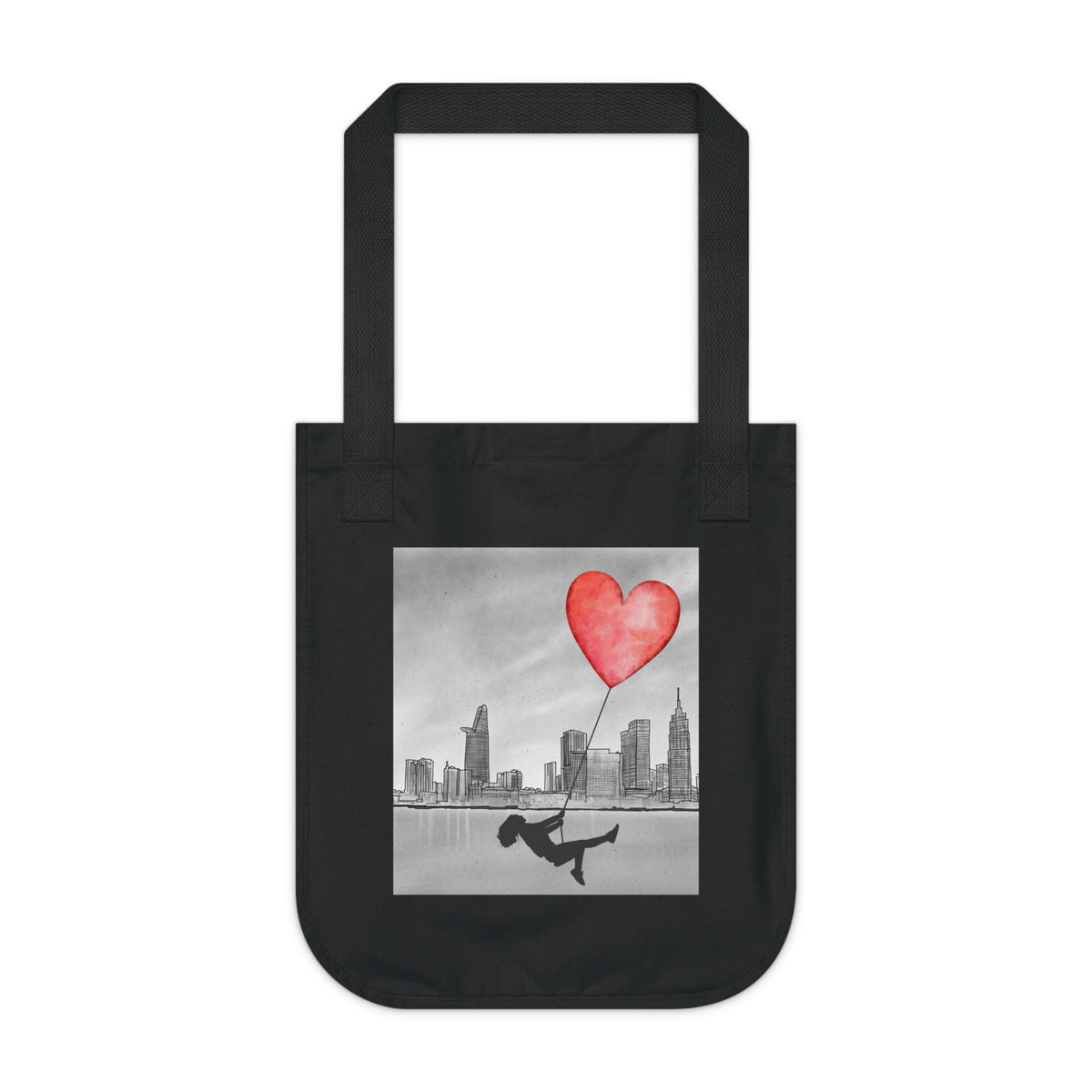 A black canvas tote bag with a square graphic print on the front. The print features a black-and-white city skyline, a person sliding on a rope, and a red heart-shaped balloon.