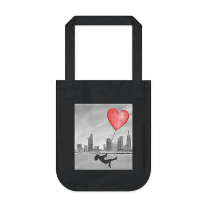 A black canvas tote bag with a square graphic print on the front. The print features a black-and-white city skyline, a person sliding on a rope, and a red heart-shaped balloon.