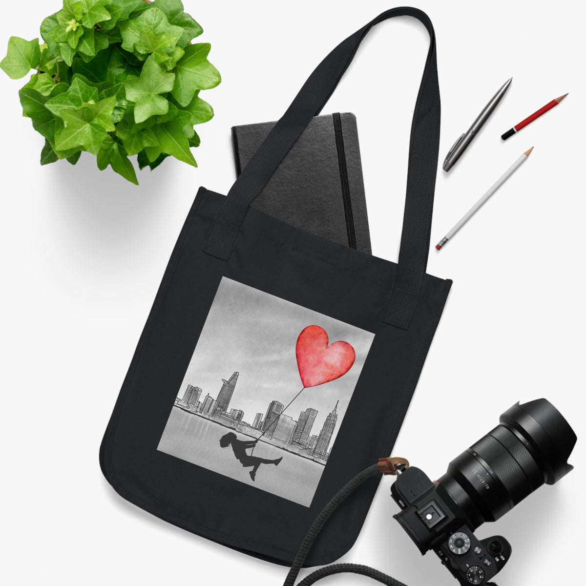 Front view of a black canvas tote bag featuring a grayscale city skyline and a silhouetted person swinging from a red heart-shaped balloon, with the heart as the only colored element in the design. the tote is styled on a white background with a notebook, camera, pens, and a green plant.